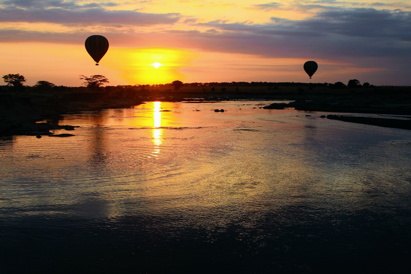 An image of a lake during sunset with hot air balloons rising in the background.